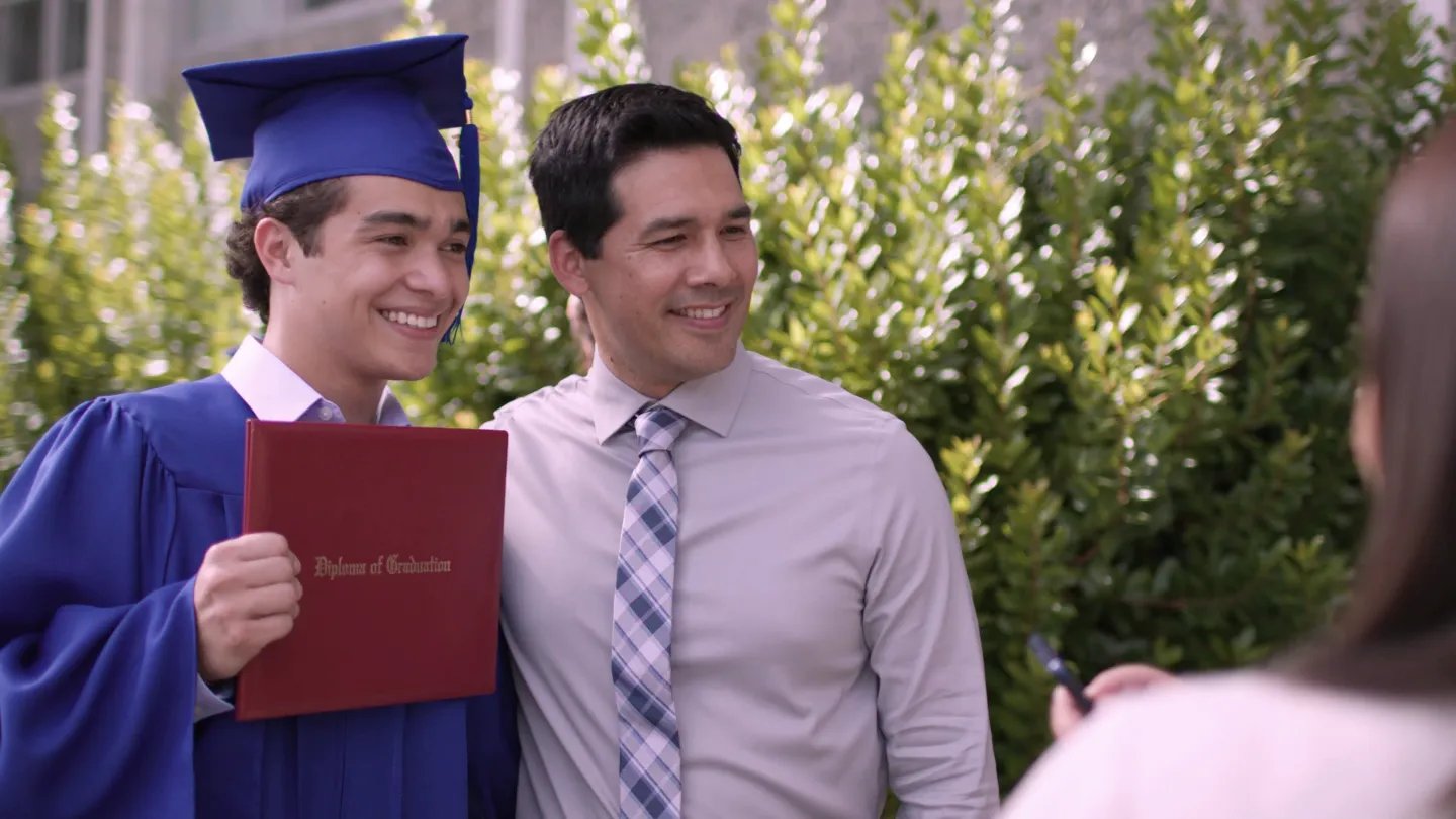 A student in a cap and gown shows off his diploma while standing next to his proud teacher.